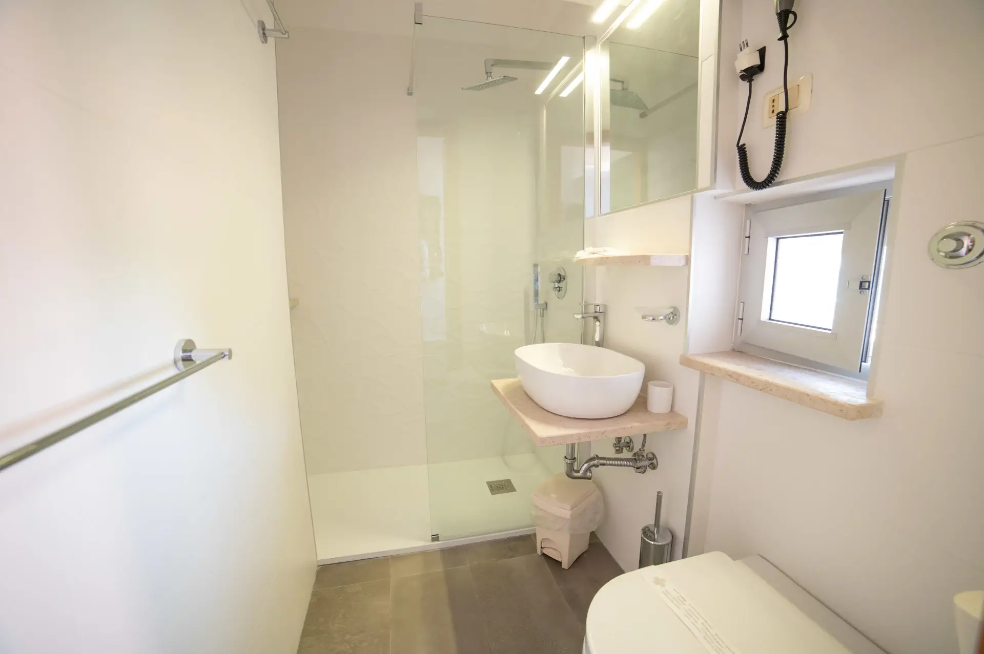 Bagno luminoso con ampia doccia in vetro, lavabo da appoggio e finestra.