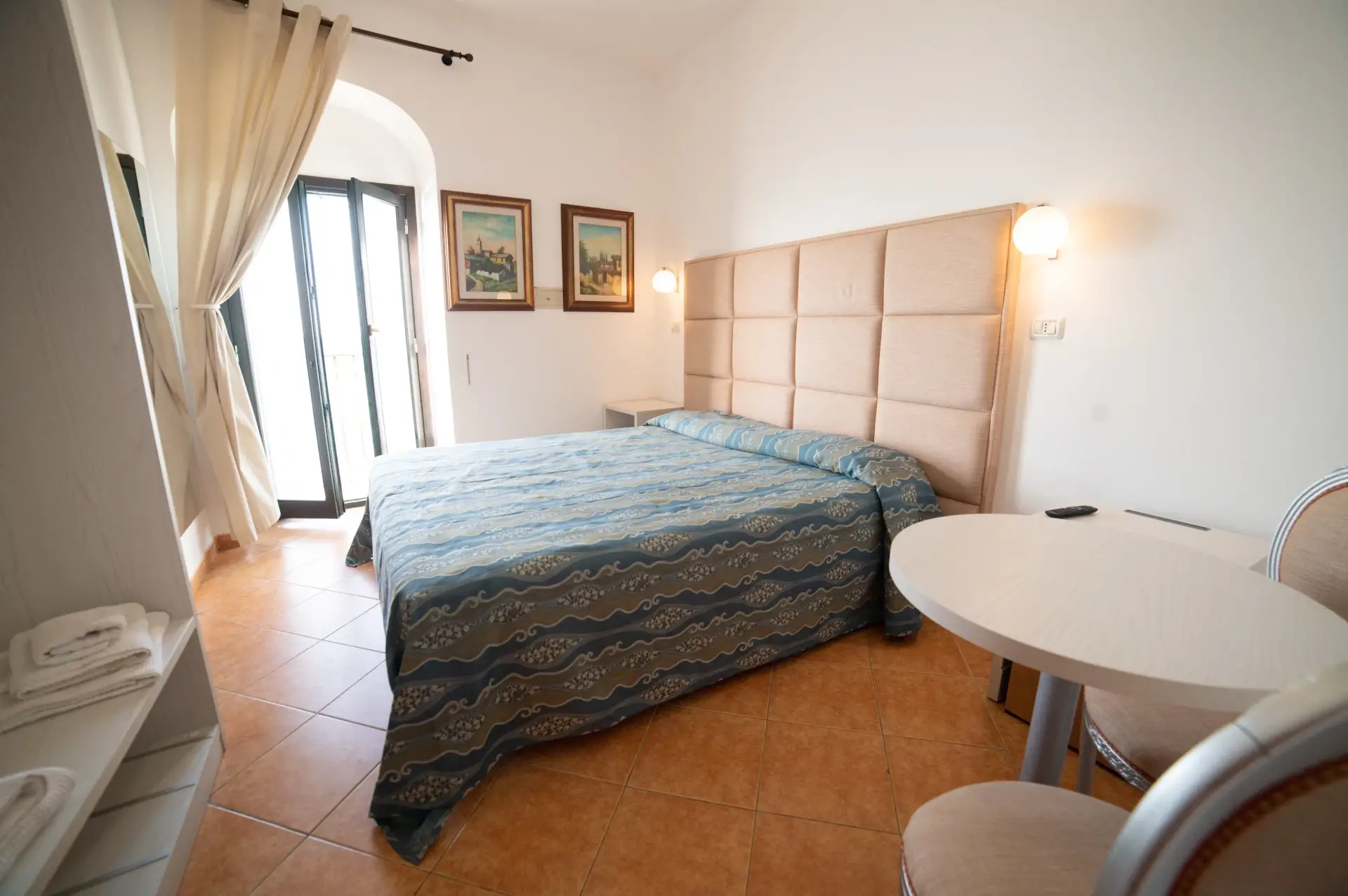 Camera matrimoniale con letto, testiera imbottita, tavolino e portafinestra.